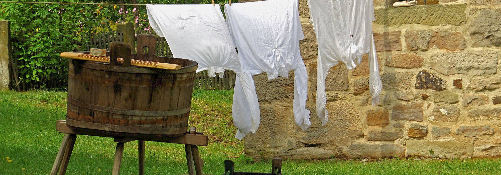Auf einer Wiese steht ein großer Holztrog mit Löffel auf einem Hocker, auf dem Boden zwei kleinere Tröge. Im Hintergrund trocknen drei weiße Hemden auf einer Wäscheleine an einem Haus