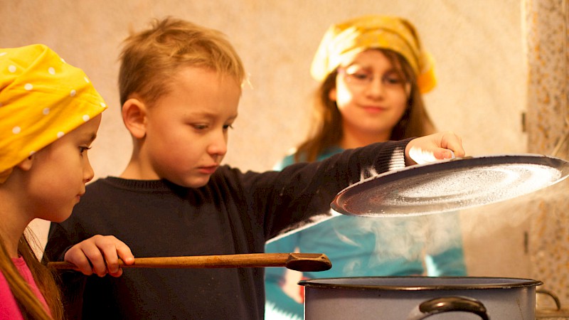 Drei Kinder im Grundschulalter stehen um einen großen Topf herum. Das Kind in der Mitte hebt vorsichtig den Deckel, aus dem Dampf aufsteigt.