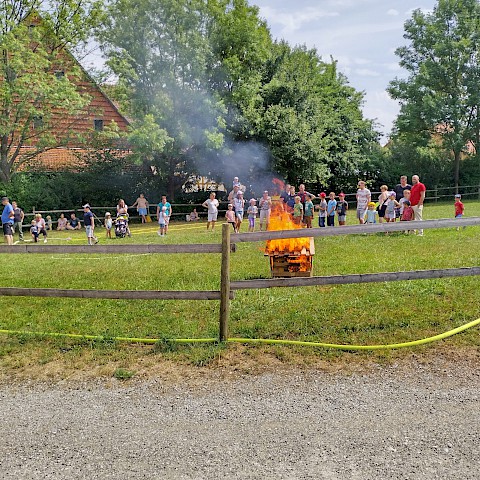 Brennende Miniatur-Hütte auf der Koppel, im Hintergrund hinter dem Zaun etwa 20 Zuschauende.