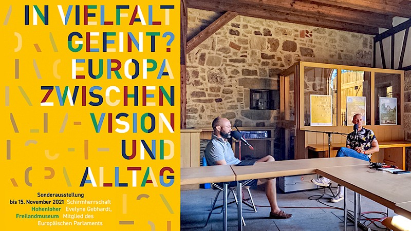 Ausstellungsplakat "In Vielfalt geeint?" im linken Bilddrittel, daneben sizten sich Stas Davydchyk, Denis Zipa und Silas Fürstenberger in einem Fachwerkgebäude gegenüber und sprechen in ihre Mikrofone