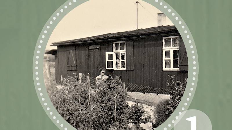 "Themenreihe Wohnbaracke aus Gschlachtenbretzingen" 1. Historische Schwarz-Weiß-Fotografie einer alten Baracke als Wohnhaus umfunktioniert. Aus dem Fenster schaut eine Person heraus, vor dem Gebäude steht eine Frau am Zaun, welcher das Gelände eingrenzt